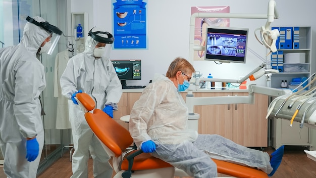 Nurse with protection suit inviting next patient in stomatology room for teeth examination during coronavirus. Assistant and dentist doctor wearing overall, face shield, mask, gloves in dental clinic