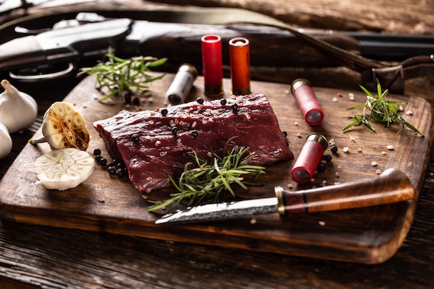 Photo nutrient rich raw deer venison prepared for a cooking process on a rustic wooden desk with roasted garlic, rosemary and huntig accesories like shot gun and ammunition.