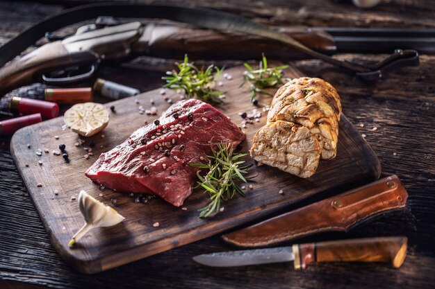 Photo nutrient rich raw deer venison prepared for a cooking process on a rustic wooden desk with roasted garlic, rosemary and huntig accesories like shot gun and ammunition