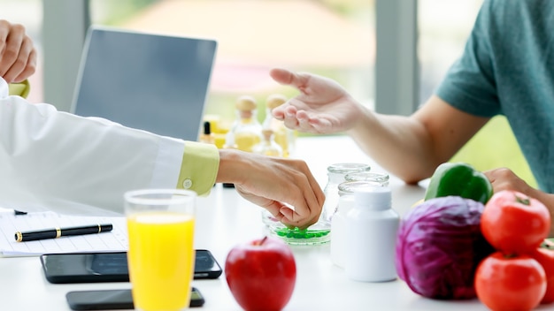 영양사의 손이 환자에게 보여주기 위해 탁자 위의 병에서 녹색 알약을 움켜쥐고 있습니다. 과일과 야채, 주스, 노트북, 영양사 사무실 테이블에 휴대 전화.