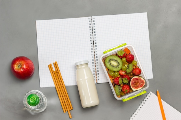 Nutritious lunch box of fruits on open notebook with milk bottle, apple, bottle of water and pencils