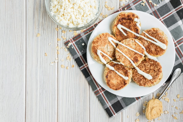 Frittelle di ricotta di avena con panna acida e burro di arachidi su fondo di legno bianco.