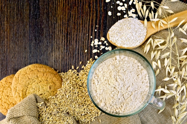 Farina d'avena in una ciotola di vetro, fiocchi di crusca in un cucchiaio, gambi di avena e biscotti di farina d'avena sul saccheggio su uno sfondo di assi di legno