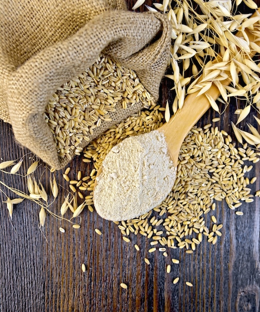 Oat flour in a wooden spoon, a bag of oats and oat stalks on a dark wooden board on top