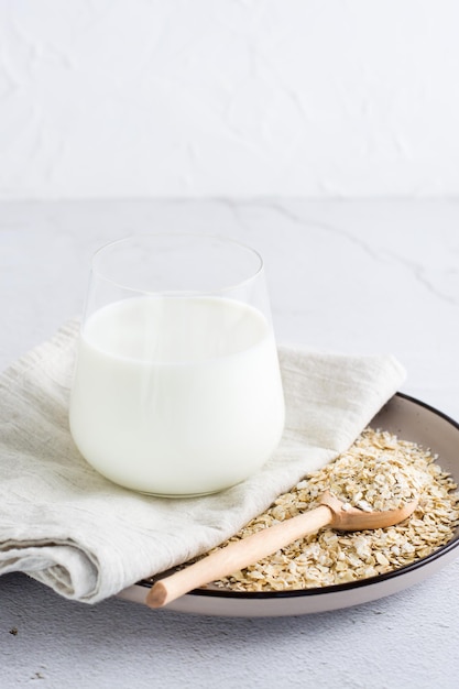 Oat milk in a glass on a plate with dry oatmeal on a light table Alternative plant food Vertical view