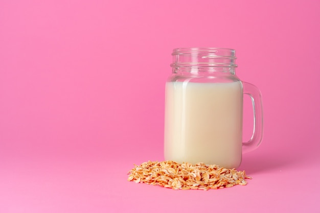 Oat milk in glassware and dry oat flakes on pink close up