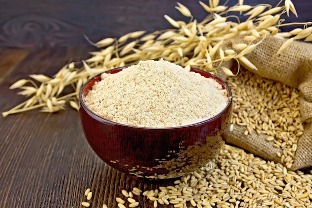 Oaten flour in a bowl, bag with oat and stalks of oats on the background of dark wood planks