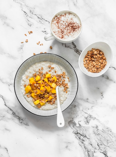 Photo oatmeal coconut milk gluten free porridge with fresh mango crispy granola and cappuccino on a light marble background top view