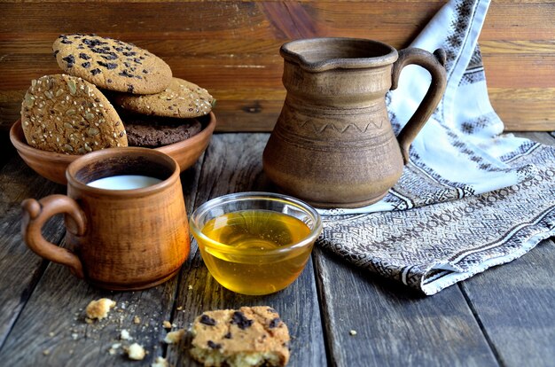 Oatmeal cookies in a ceramic plate, milk in a ceramic mug, earthenware jug, napkin on old wooden boards.