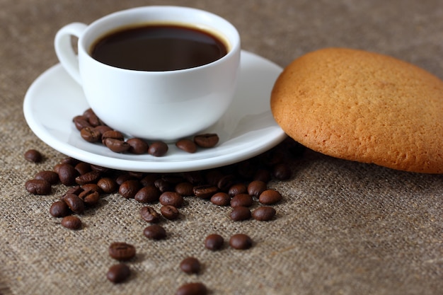 Овсяное печенье, кружка с кофе, блюдце и кофейные зерна, разбросанные на мешковину скатертью.