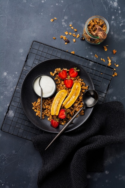 Oatmeal granola with yoghurt, fresh strawberries and banana, chia seeds, sunflower and honey in black ceramic plate  . Top view.