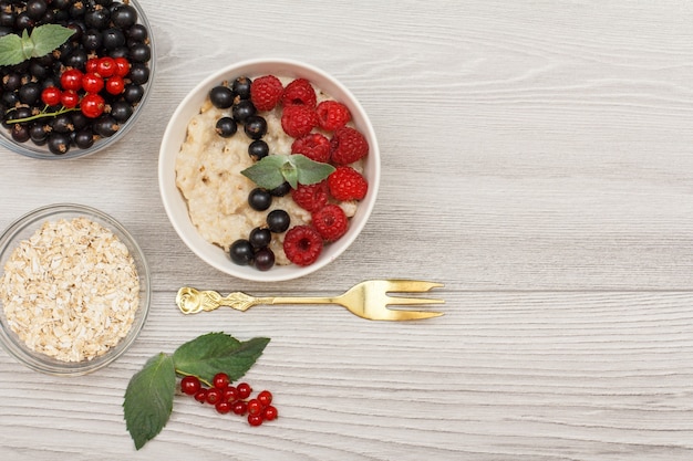 Oatmeal porridge in porcelain bowl with currant berries and raspberries, decorated with mint leaves. Glass bowls with oat flakes and currant berries. Top view with copy space