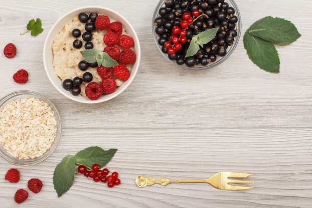 Oatmeal porridge in porcelain bowl with currant berries and raspberries, decorated with mint leaves. Glass bowls with oat flakes and currant berries with metal fork. Top view with copy space