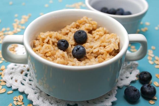 Oatmeal porridge with fresh blueberries and oat flakes in white bowl on blue  with white rustic knitted napkin close up. Selective soft focus. . Text copy space.