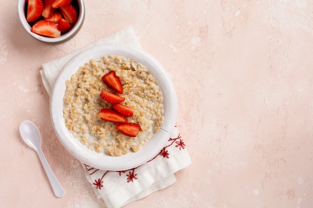 Oatmeal porridge with strawberries in a white plate on a linen napkin on pink surface. Top view.