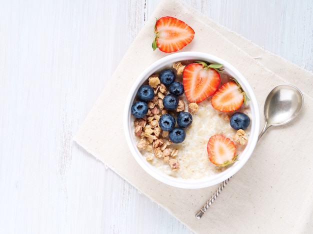 Oatmeal porridge with strawberry, blueberry, granola on white wooden background. 