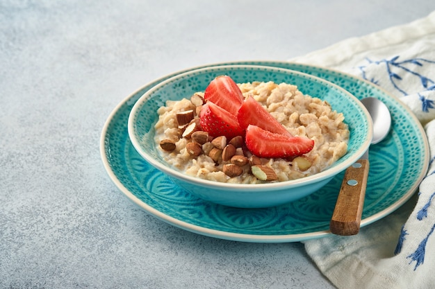 Oatmeal porridge with strawberry slices, nuts almonds and honey in blue bowl on grey table. Healthy eating, dieting, vegetarian food concept. Place for text.