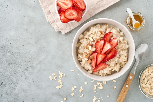 Oatmeal porridge with strawberry slices, nuts almonds and honey in bowl on grey table. Healthy eating, dieting, vegetarian food concept. Place for text