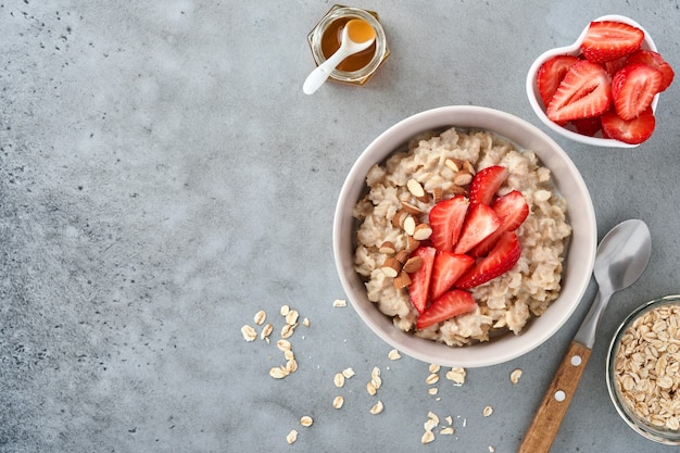 Oatmeal porridge with strawberry slices, nuts almonds and honey in bowl on grey table. Healthy eating, dieting, vegetarian food concept. Place for text.