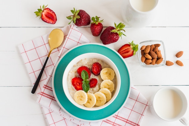 Oatmeal with strawberries and banana slices in a white plate standing on a white wooden table.