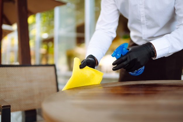 Ober draagt beschermend gezichtsmasker en handschoenen tijdens het desinfecteren van tafels op terras.