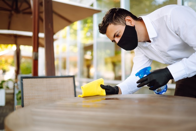 Ober draagt beschermend gezichtsmasker en handschoenen tijdens het desinfecteren van tafels op terras.