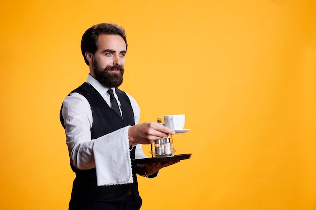 Ober geeft een kopje koffie aan de klant voor de camera en doet alsof hij mensen bedient in een gastronomisch restaurant. Geschoolde stijlvolle man in formele kledij met handdoek over arm en voedselschotel.