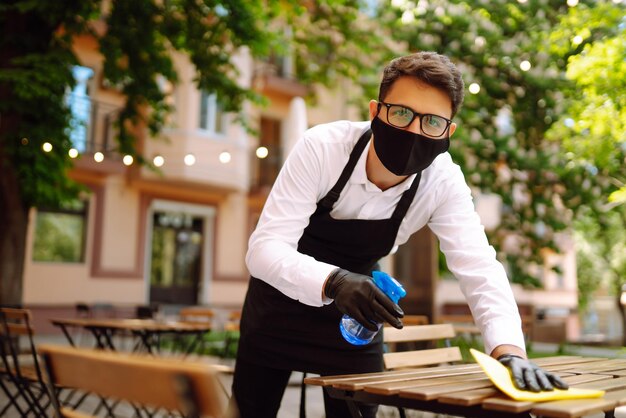 Ober met beschermend gezichtsmasker en handschoenen die de restauranttafel schoonmaken voor de volgende klant