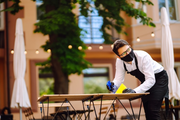 Ober met beschermend masker en handschoenen die de restauranttafel desinfecteren voor de volgende klant.