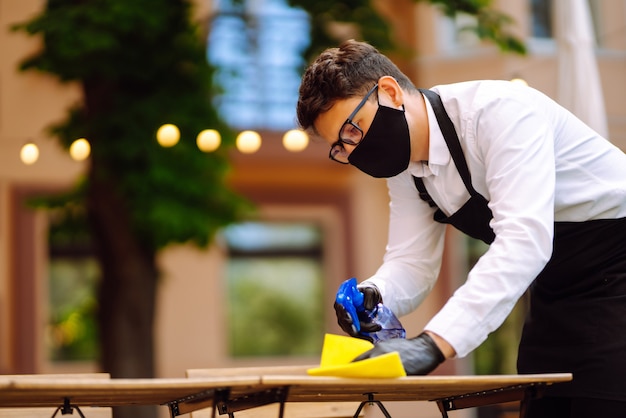 Ober met beschermend masker en handschoenen die de restauranttafel desinfecteren voor de volgende klant.