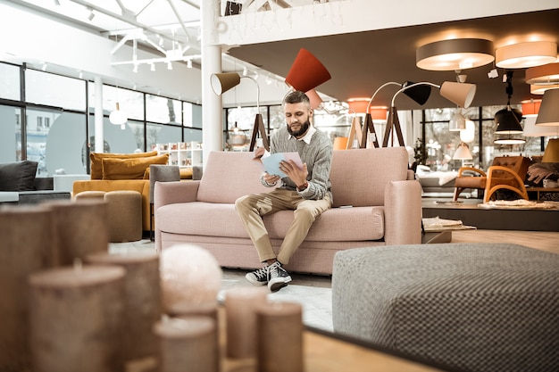 Observing magazine. Curious bearded man observing catalog with variants of furniture while sitting in the middle of furniture store