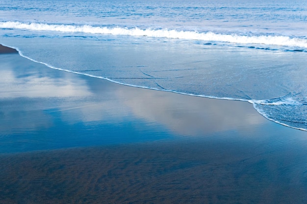 Oceaanstrand met zwart vulkanisch zand de lucht wordt weerspiegeld in de teruggerolde golf van de branding