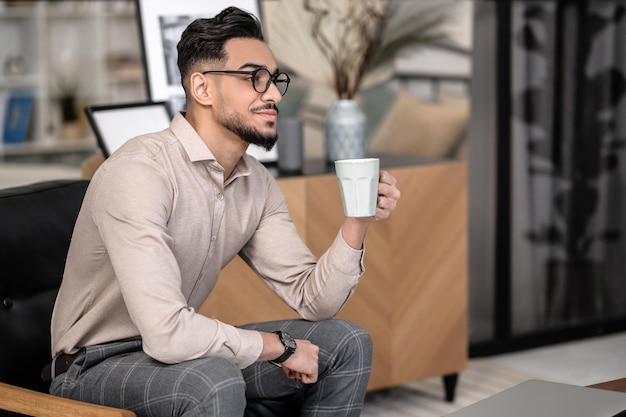 Ochtend koffie. Mediteren jonge, bebaarde stijlvolle man met koffie zijwaarts zittend naar de camera in de kamer thuis