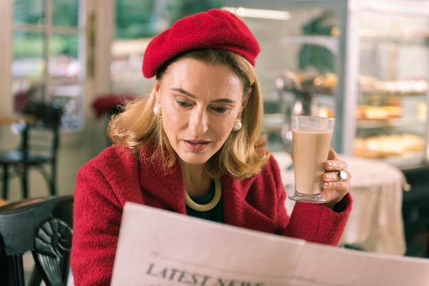 Ochtendkrant. Rijpe Franse elegante stijlvolle vrouw lezing ochtendkrant zitten in bakkerij
