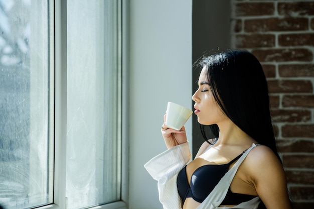 Ochtendritueel Meisje sexy borsten koffie drinken in de buurt van vensterbank Aantrekkelijke vrouwelijke lingerie ontspannen thuis Perfecte koffietijd Maar eerst koffie Rust en vrede Sip van romige cappuccino