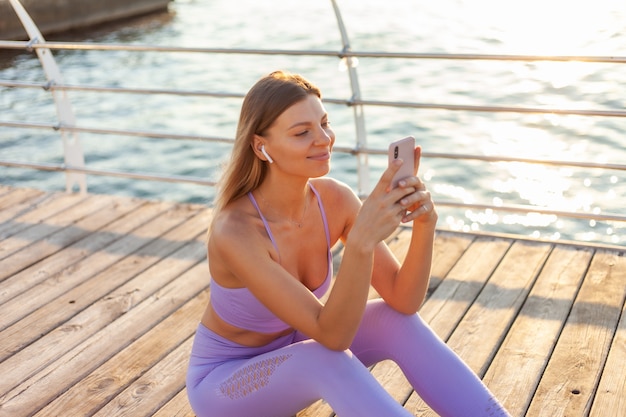 Ochtendtraining. De jonge sportvrouw in sportkleding gebruikt smartphone terwijl het zitten op het strand bij zonsopgang