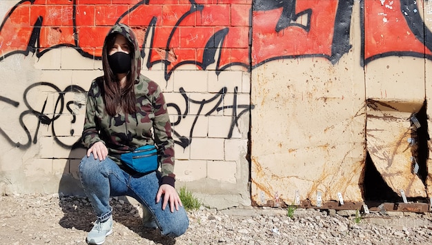 Oekraïne, Kiev - 24 april 2020. Een mooie vrouw met een capuchon en een beschermend masker, gekleed in een spijkerbroek en een kaki camouflagejasje, zit bij een muur met graffiti. Op straat.