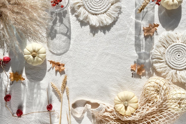 Off white natural Fall decorations with copy-space. Flat lay on white textile, place for text. Pumpkins, dry leaves and rowan berries, macrame pads, wattle leaves and wheat ears. Sunlight, shadows .