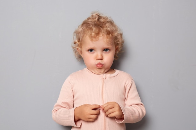 Offended sad baby girl with blonde wavy hair wearing rose shirt isolated over gray background looking at camera with blowing cheeks having bad mood