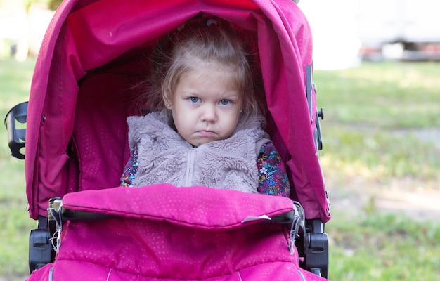 Offended and sad little girl sitting in stroller in summer park