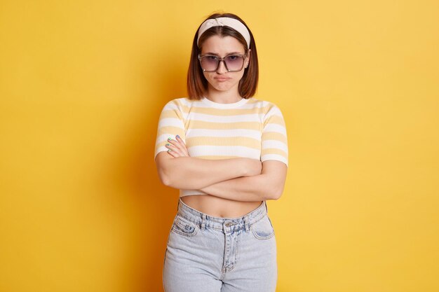 Offended unhappy woman wearing striped Tshirt hair band and sunglasses standing isolated over yellow background standing folded hands expressing sorrow and sadness