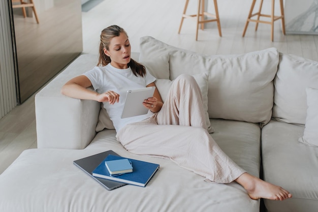 Offended young Hispanic woman in white tshirt and pants laying on cozy couch with laptop and books uses tablet makes video call with sad face expression Tired at distant lesson via internet at home