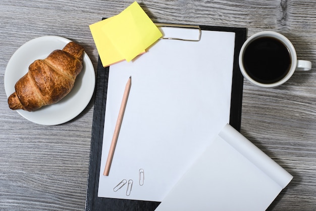 Office accessorise on wooden gray table: clipboard, cup of coffee, fresh croissant