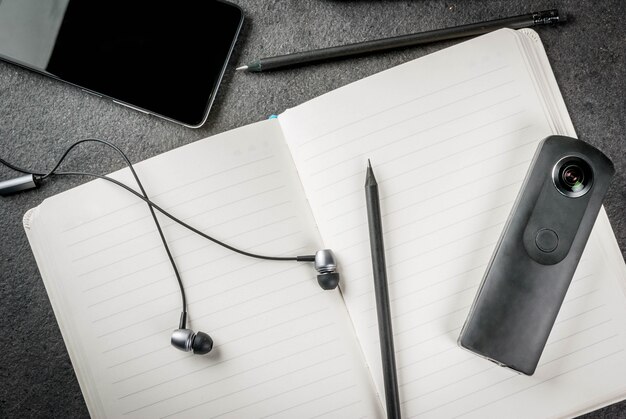 Office, black desktop with appliances. . Notepad, panoramic camera pens (pencils), smartphone and headphones on the table.