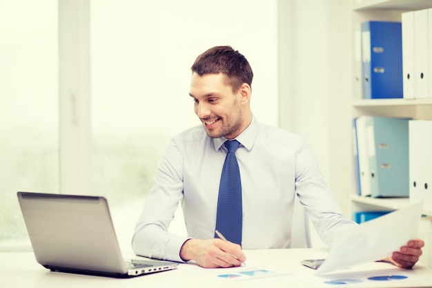 office, business, technology, finances and internet concept - smiling businessman with laptop computer and documents at office