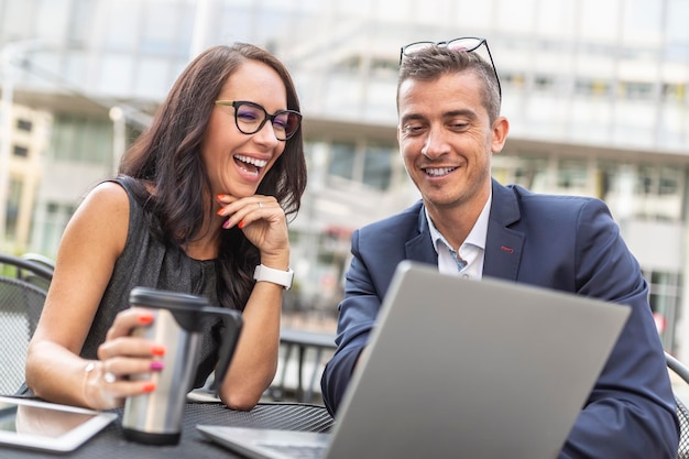 Office colleagues discuss and laugh over a laptop sitting with a coffee outdside