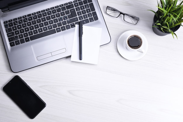 ラップトップ、スマートフォン、一杯のコーヒーと消耗品、木製の背景の上のオフィスの机のテーブル。コピースペース、フラットレイの上面図。