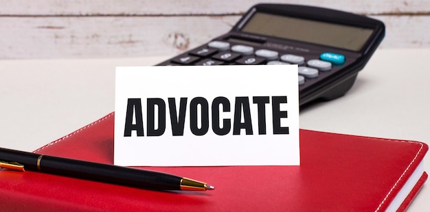 On the office desk there is a burgundy notebook a calculator a pen and a white card with the text ADVOCATE Business concept