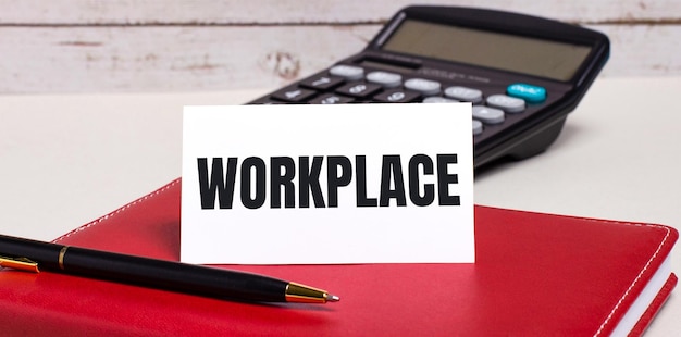 На офисном столе есть бордовый блокнот, калькулятор, ручка и белая карточка с текстом WORKPLACE Business concept
