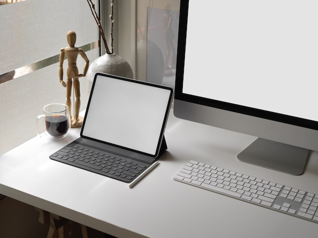 Office desk with blank screen tablet, computer device, coffee cup and decorations beside window